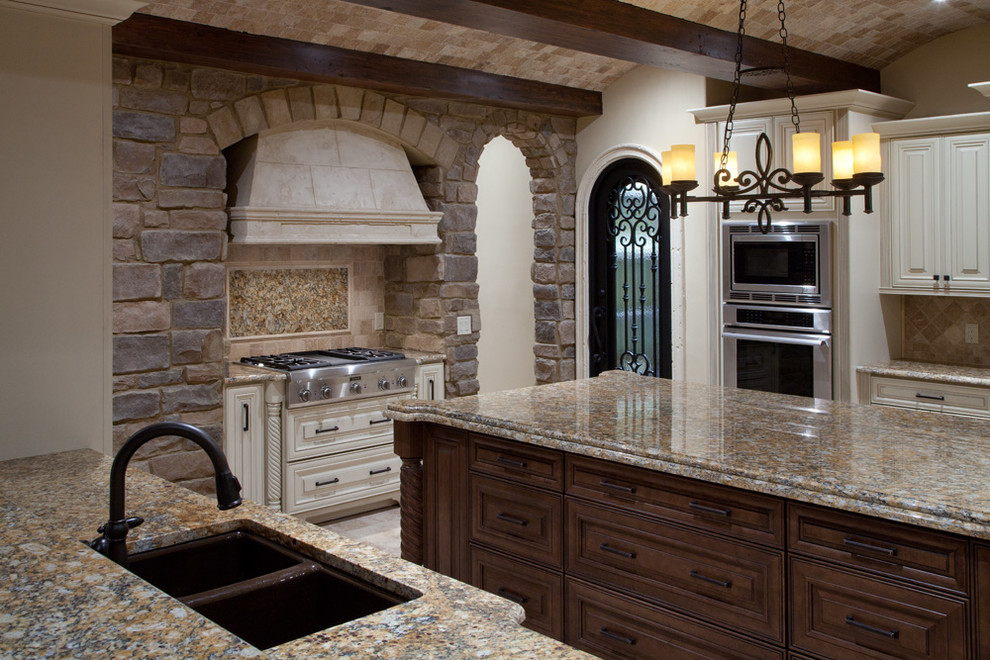 This is an example of a mid-sized mediterranean u-shaped eat-in kitchen in Orlando with a double-bowl sink, raised-panel cabinets, dark wood cabinets, granite benchtops, beige splashback, stone tile splashback, stainless steel appliances, travertine floors and with island.