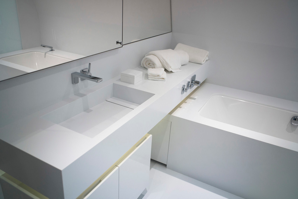 Design ideas for a mid-sized contemporary master bathroom in London with flat-panel cabinets, a freestanding tub, a wall-mount toilet, white tile, white cabinets, mirror tile, white walls, vinyl floors, an integrated sink and solid surface benchtops.