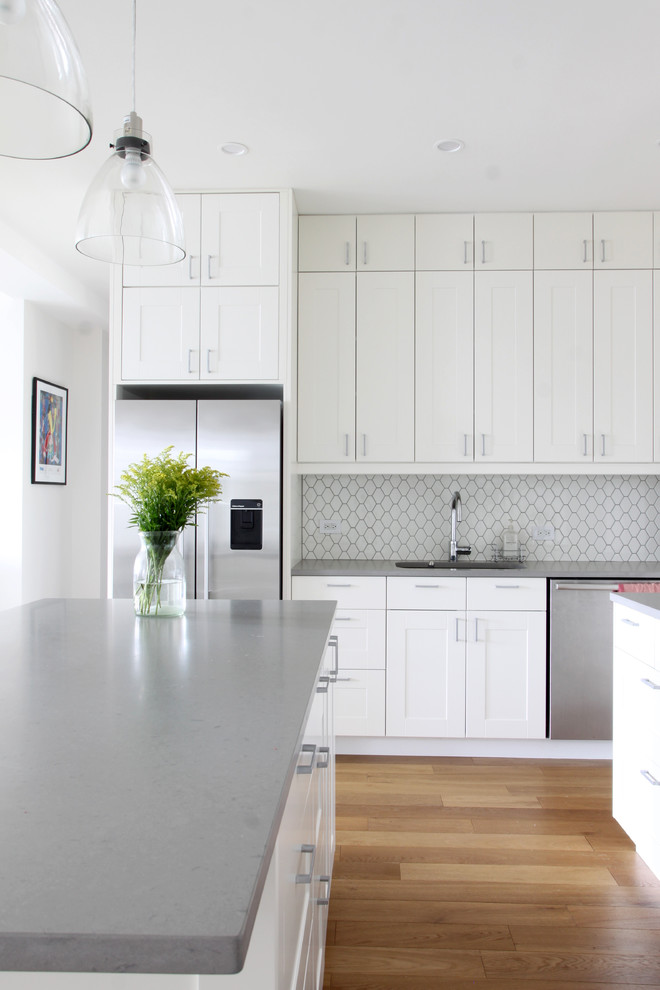 This is an example of a mid-sized transitional l-shaped kitchen in New York with shaker cabinets, white cabinets, quartz benchtops, white splashback, ceramic splashback, stainless steel appliances, medium hardwood floors and multiple islands.