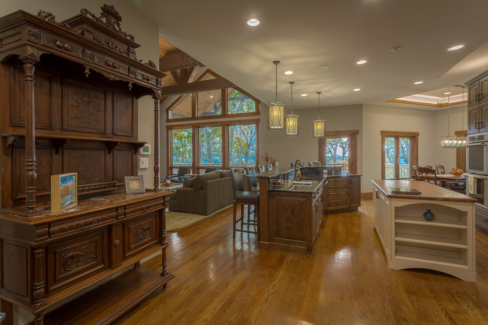 This is an example of a large country l-shaped open plan kitchen in Charlotte with raised-panel cabinets, dark wood cabinets, granite benchtops, stone slab splashback, stainless steel appliances, medium hardwood floors, multiple islands, a double-bowl sink, beige splashback and brown floor.