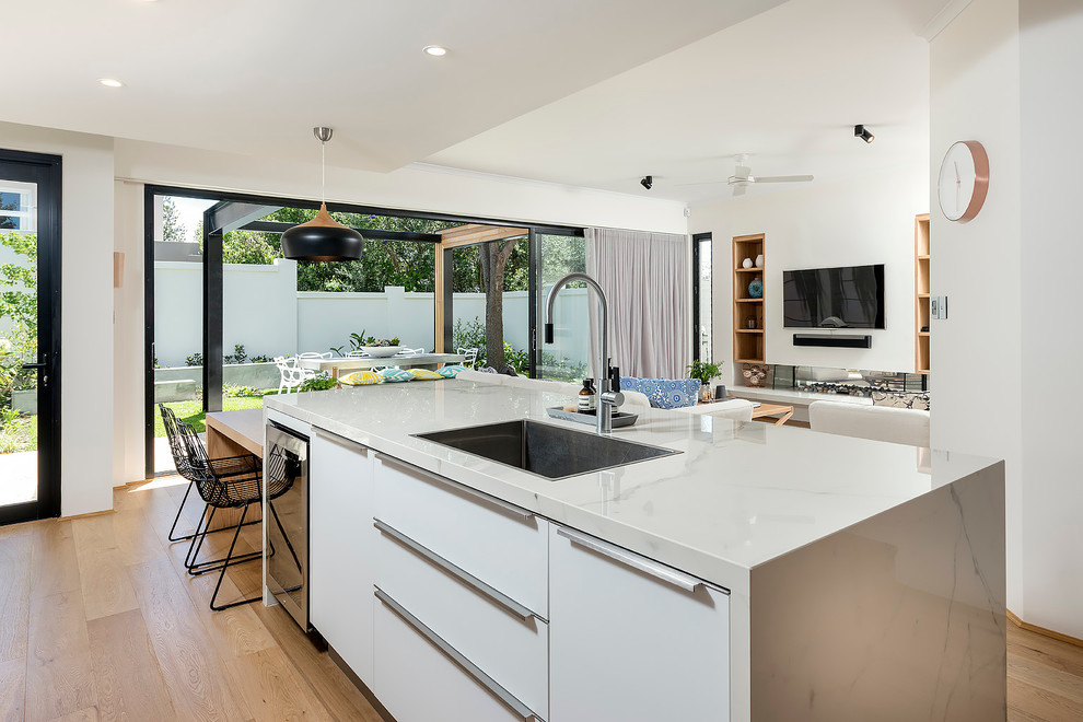 Design ideas for a large modern single-wall open plan kitchen in Perth with a double-bowl sink, flat-panel cabinets, white cabinets, tile countertops, metallic splashback, mirror splashback, black appliances, light hardwood flooring, an island, brown floors and white worktops.