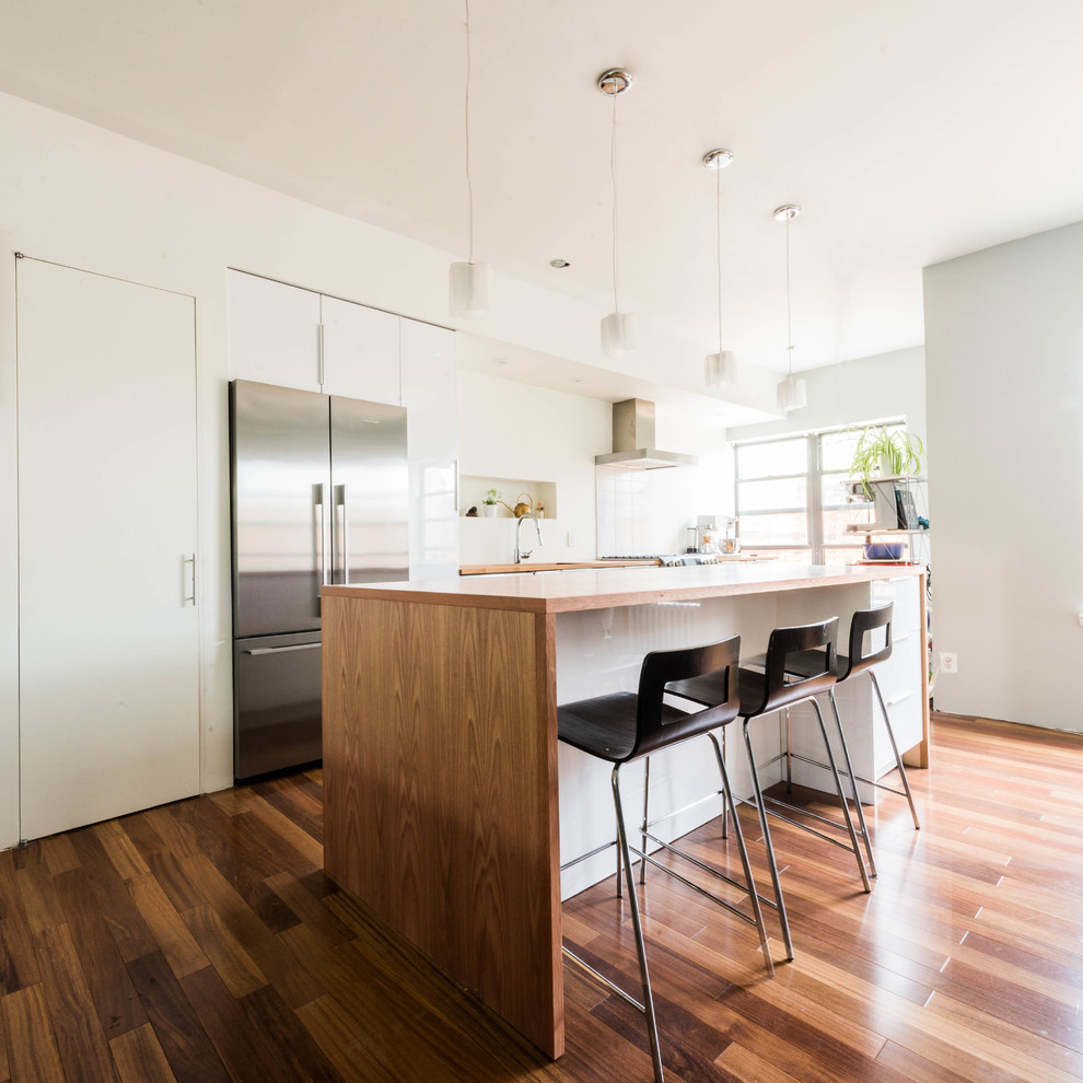 This is an example of a mid-sized contemporary l-shaped eat-in kitchen in New York with flat-panel cabinets, white cabinets, wood benchtops, white splashback, stone slab splashback, stainless steel appliances, medium hardwood floors and with island.