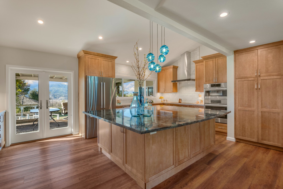 Example of a large classic u-shaped medium tone wood floor, brown floor and vaulted ceiling open concept kitchen design in San Francisco with an undermount sink, shaker cabinets, medium tone wood cabinets, solid surface countertops, white backsplash, ceramic backsplash, stainless steel appliances, an island and blue countertops