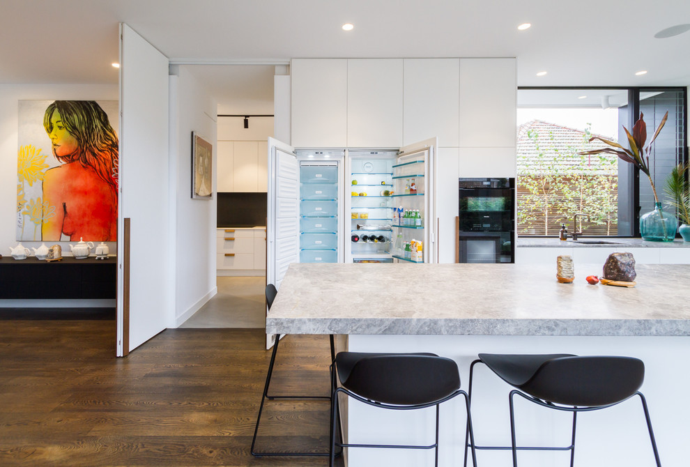 This is an example of a large modern l-shaped open plan kitchen in Melbourne with an undermount sink, flat-panel cabinets, white cabinets, marble benchtops, grey splashback, black appliances, medium hardwood floors, with island, brown floor, grey benchtop and limestone splashback.