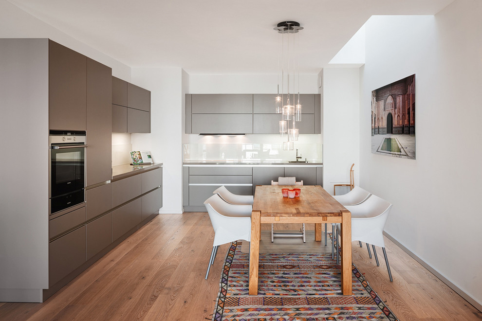 Design ideas for a large contemporary l-shaped eat-in kitchen in Berlin with a single-bowl sink, flat-panel cabinets, grey cabinets, white splashback, glass sheet splashback, panelled appliances, medium hardwood floors and no island.