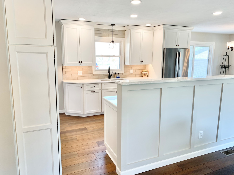 Photo of a mid-sized transitional l-shaped open plan kitchen in Other with an undermount sink, flat-panel cabinets, white cabinets, quartz benchtops, beige splashback, ceramic splashback, stainless steel appliances, vinyl floors, with island, brown floor and white benchtop.