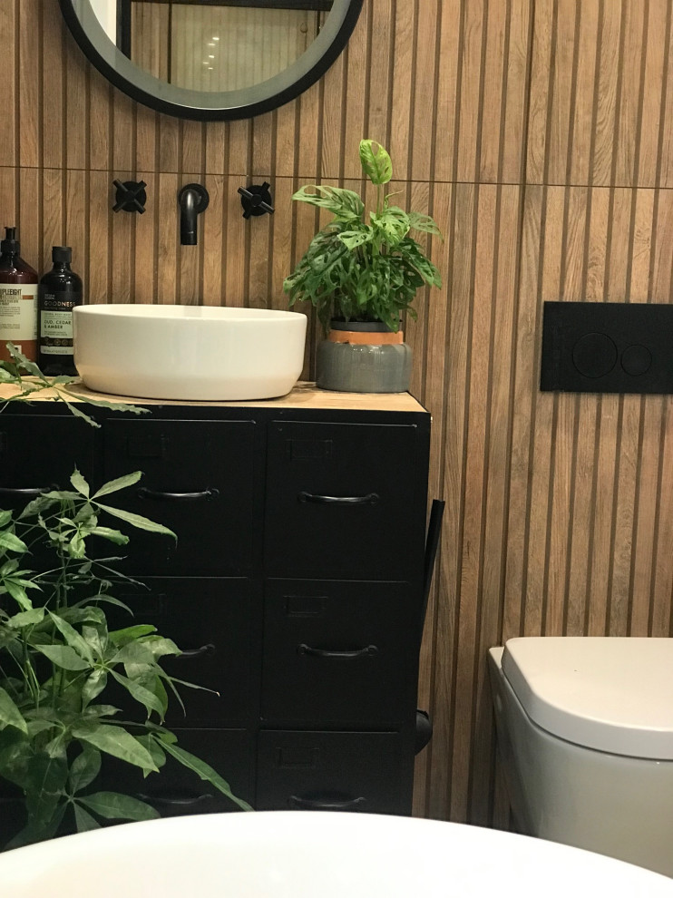 This is an example of a mid-sized tropical master bathroom in Sussex with furniture-like cabinets, black cabinets, a freestanding tub, a curbless shower, a wall-mount toilet, black and white tile, wood-look tile, multi-coloured walls, limestone floors, a pedestal sink, wood benchtops, beige floor, a sliding shower screen, brown benchtops, a single vanity and a freestanding vanity.
