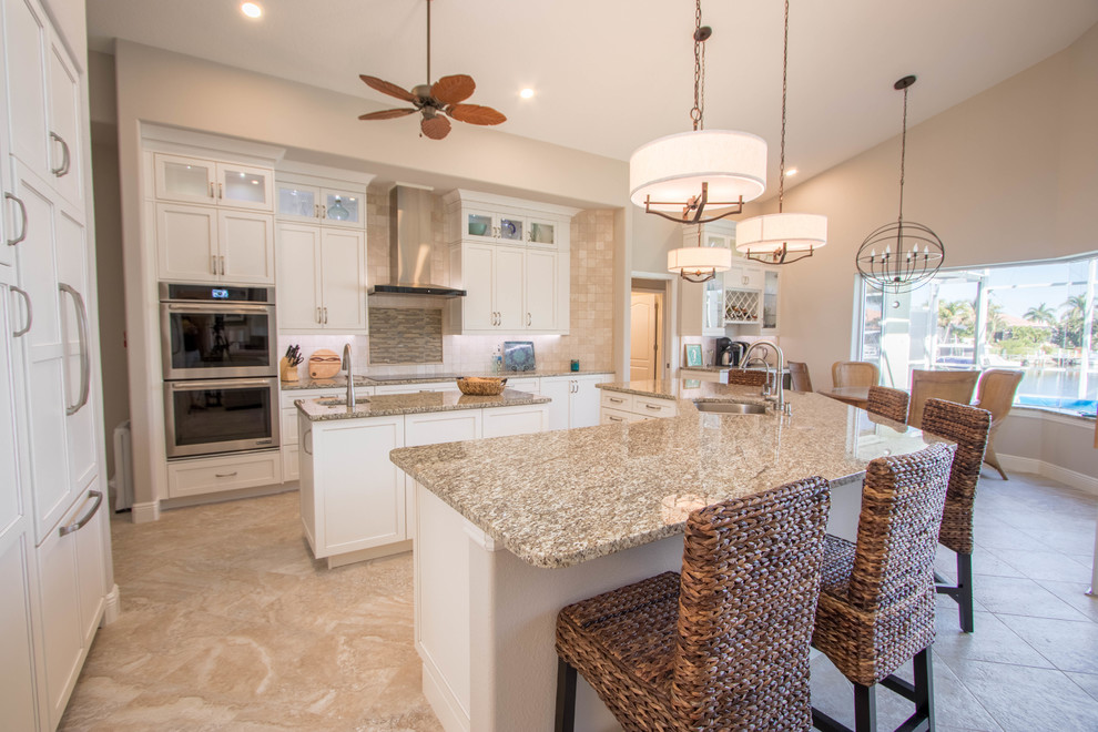 Inspiration for an expansive transitional l-shaped open plan kitchen in Miami with a double-bowl sink, shaker cabinets, white cabinets, granite benchtops, beige splashback, stone tile splashback, stainless steel appliances, travertine floors and multiple islands.