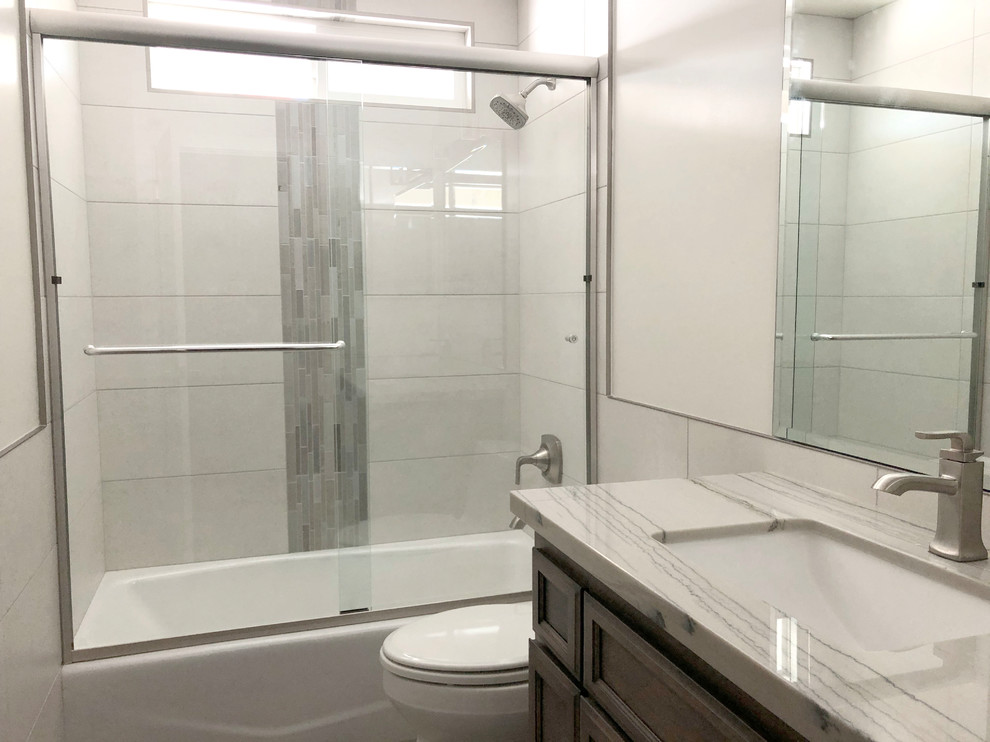 This is an example of a mid-sized transitional bathroom in Los Angeles with raised-panel cabinets, grey cabinets, an alcove tub, a shower/bathtub combo, a one-piece toilet, gray tile, porcelain tile, white walls, porcelain floors, an undermount sink, quartzite benchtops, grey floor, a sliding shower screen and grey benchtops.