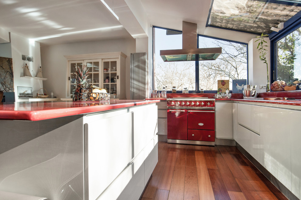 Photo of a large midcentury galley eat-in kitchen in Marseille with a double-bowl sink, flat-panel cabinets, grey cabinets, solid surface benchtops, red splashback, panelled appliances, medium hardwood floors, with island, brown floor and red benchtop.