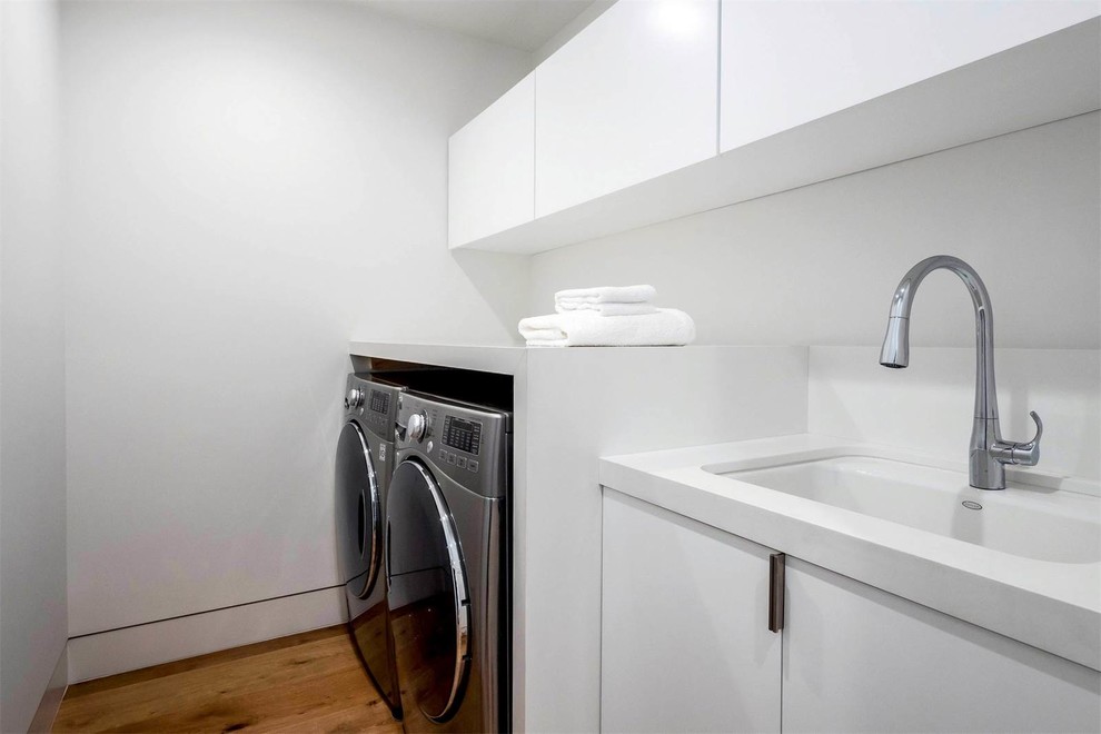 Mid-sized contemporary single-wall dedicated laundry room in Los Angeles with an undermount sink, flat-panel cabinets, white cabinets, solid surface benchtops, white walls, medium hardwood floors, a side-by-side washer and dryer and brown floor.