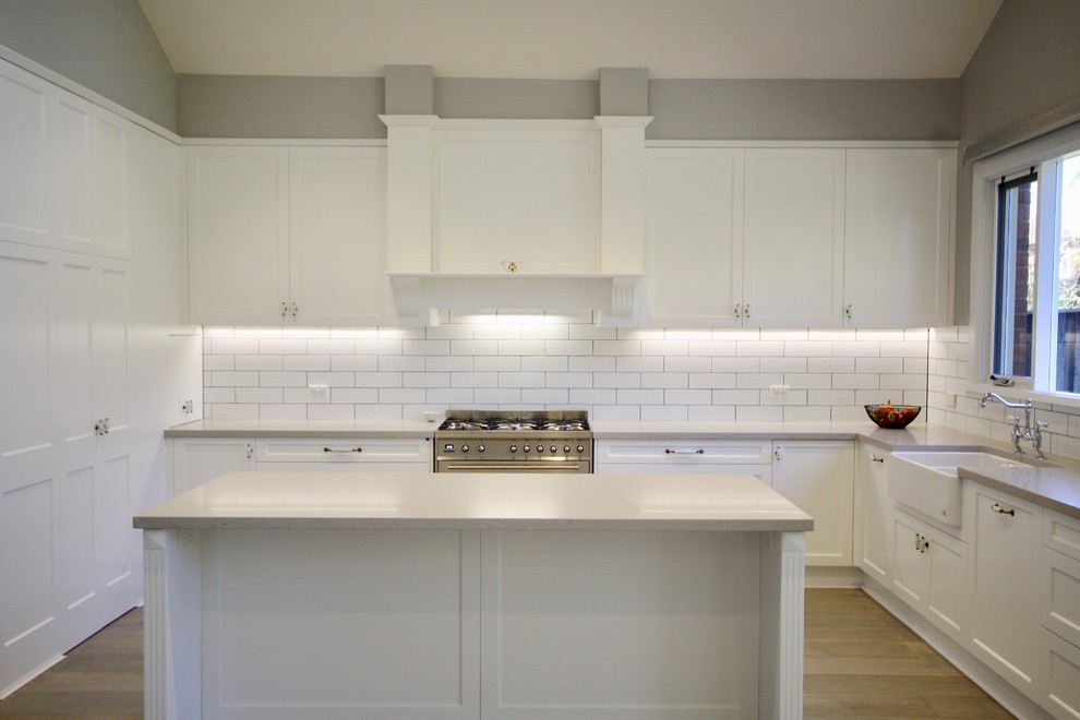 Photo of a large transitional l-shaped eat-in kitchen in Sydney with a farmhouse sink, shaker cabinets, white cabinets, quartz benchtops, white splashback, ceramic splashback, stainless steel appliances, light hardwood floors, with island and brown floor.