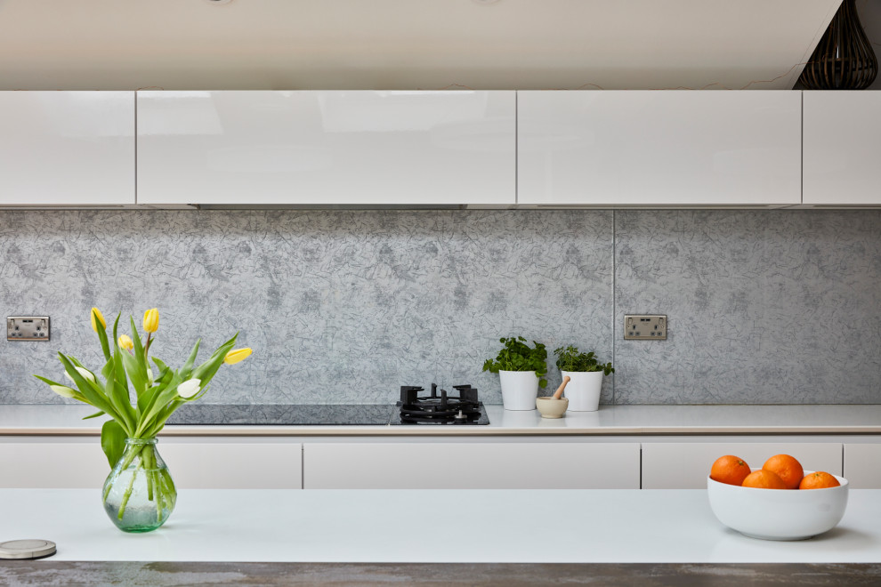 Large modern galley kitchen/diner in London with a built-in sink, flat-panel cabinets, white cabinets, granite worktops, grey splashback, metal splashback, integrated appliances, dark hardwood flooring, an island, brown floors and white worktops.
