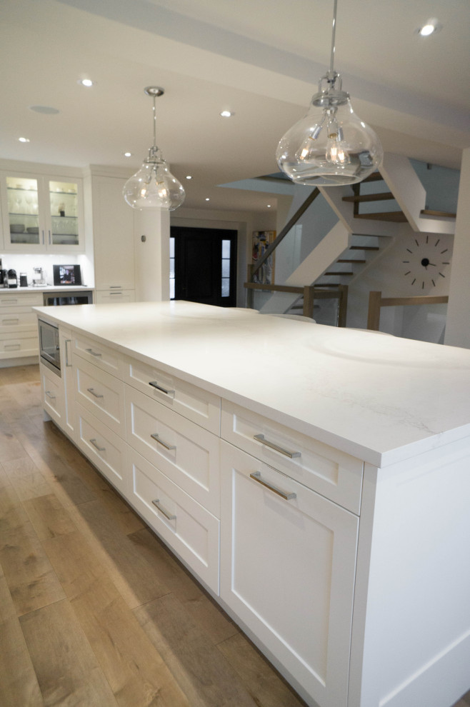 Expansive contemporary u-shaped open plan kitchen in Toronto with an undermount sink, shaker cabinets, white cabinets, quartz benchtops, grey splashback, subway tile splashback, stainless steel appliances, light hardwood floors, with island and white benchtop.
