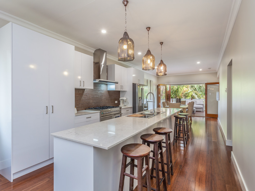 Photo of a mid-sized modern galley eat-in kitchen in Brisbane with a drop-in sink, white cabinets, quartz benchtops, metallic splashback, matchstick tile splashback, stainless steel appliances, bamboo floors and with island.