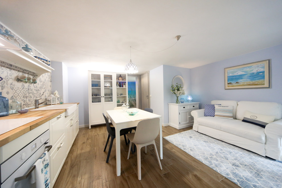 This is an example of a small beach style single-wall open plan kitchen in Other with a farmhouse sink, raised-panel cabinets, white cabinets, wood benchtops, blue splashback, porcelain splashback, white appliances, porcelain floors and no island.