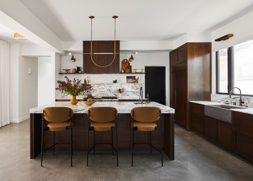 This is an example of a modern l-shaped kitchen in Orange County with a farmhouse sink, shaker cabinets, dark wood cabinets, white splashback, stone slab splashback, panelled appliances, concrete floors, with island, grey floor and white benchtop.