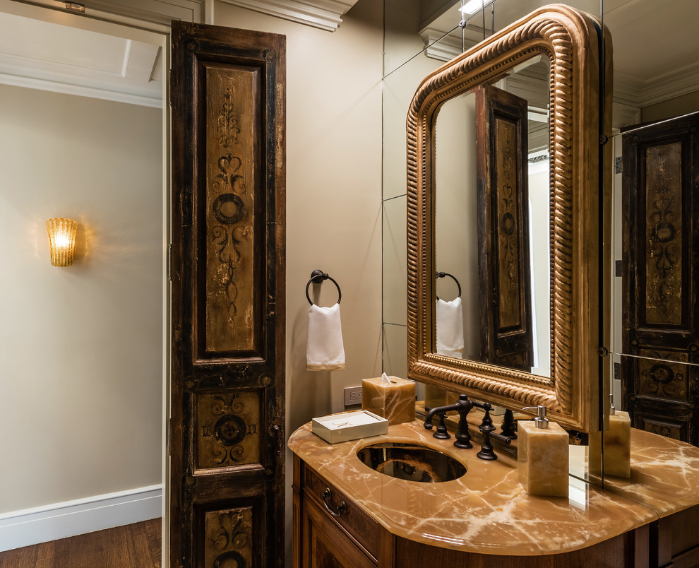 Inspiration for a mid-sized traditional powder room in Dallas with furniture-like cabinets, dark wood cabinets, a one-piece toilet, mirror tile, beige walls, porcelain floors, an undermount sink, onyx benchtops, beige floor and brown benchtops.