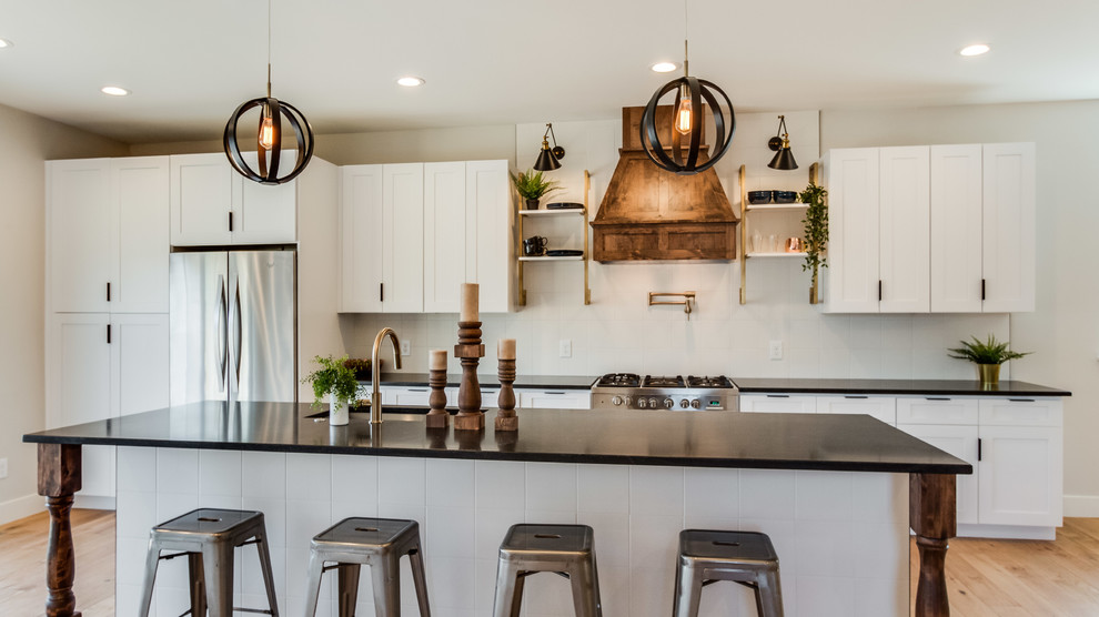 Example of a mid-sized cottage galley light wood floor and beige floor eat-in kitchen design in Denver with an undermount sink, shaker cabinets, white cabinets, white backsplash, ceramic backsplash, stainless steel appliances, an island and black countertops