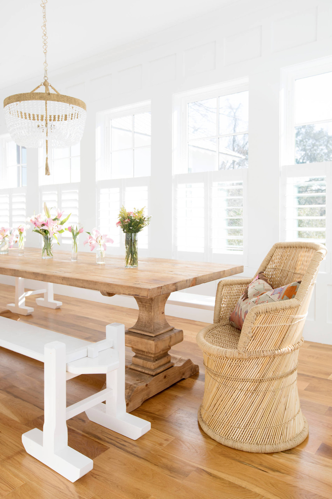 Bright dining room with a rustic wood table, white chairs, wicker chair, chandelier, and tall windows with shutters.