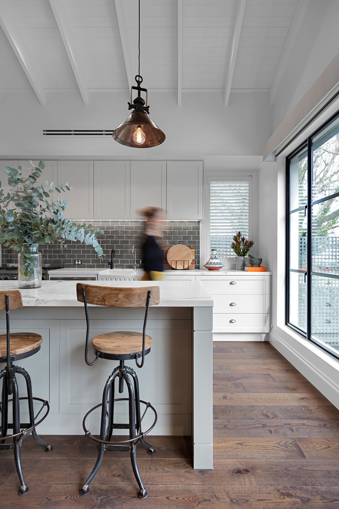 Photo of a large country l-shaped eat-in kitchen in Melbourne with a farmhouse sink, raised-panel cabinets, white cabinets, marble benchtops, grey splashback, subway tile splashback, stainless steel appliances, medium hardwood floors, with island, brown floor, white benchtop and exposed beam.