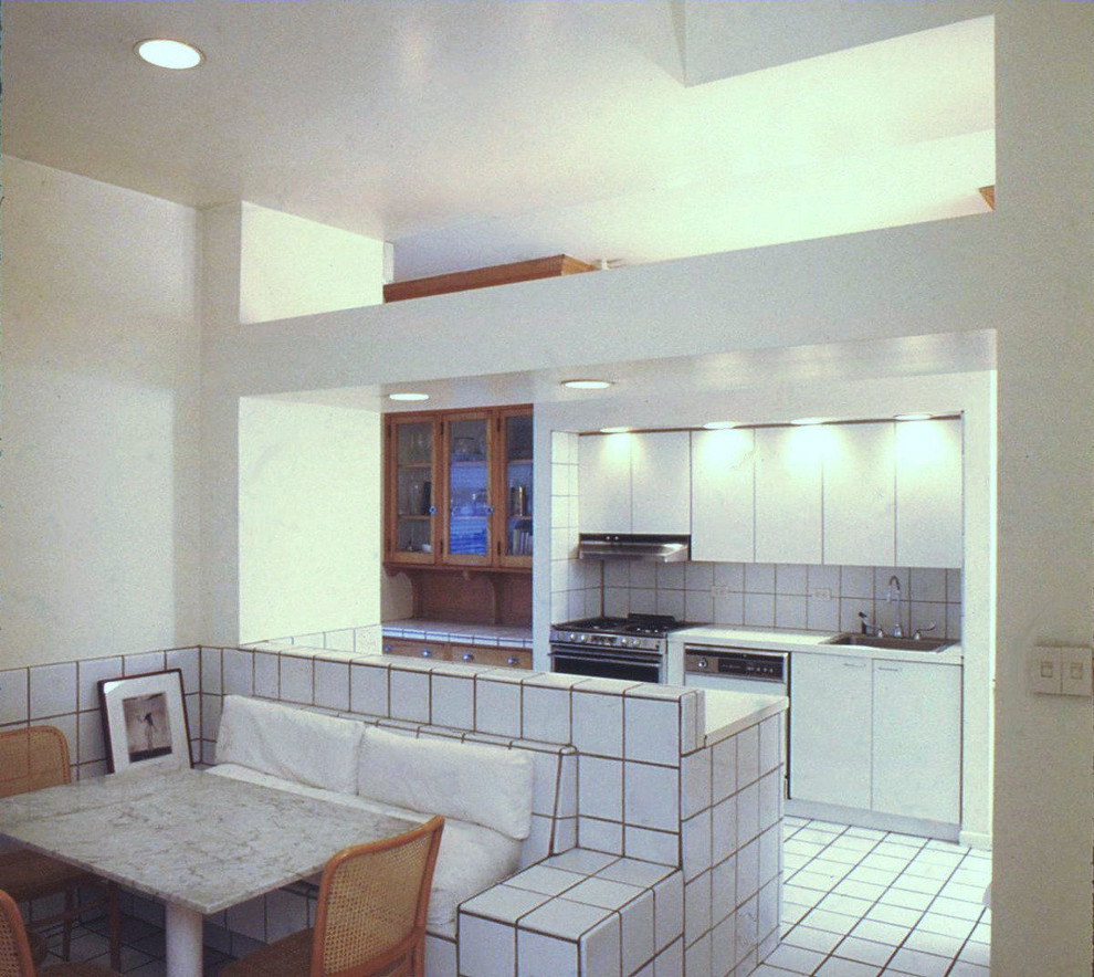 This is an example of an expansive modern u-shaped eat-in kitchen in New York with a drop-in sink, glass-front cabinets, medium wood cabinets, tile benchtops, white splashback, ceramic splashback, panelled appliances, ceramic floors and with island.