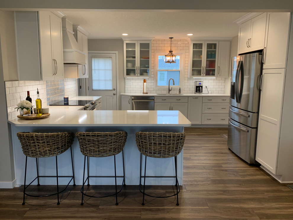 Mid-sized transitional u-shaped vinyl floor and brown floor open concept kitchen photo with an undermount sink, shaker cabinets, white cabinets, quartz countertops, white backsplash, stainless steel appliances, a peninsula and white countertops