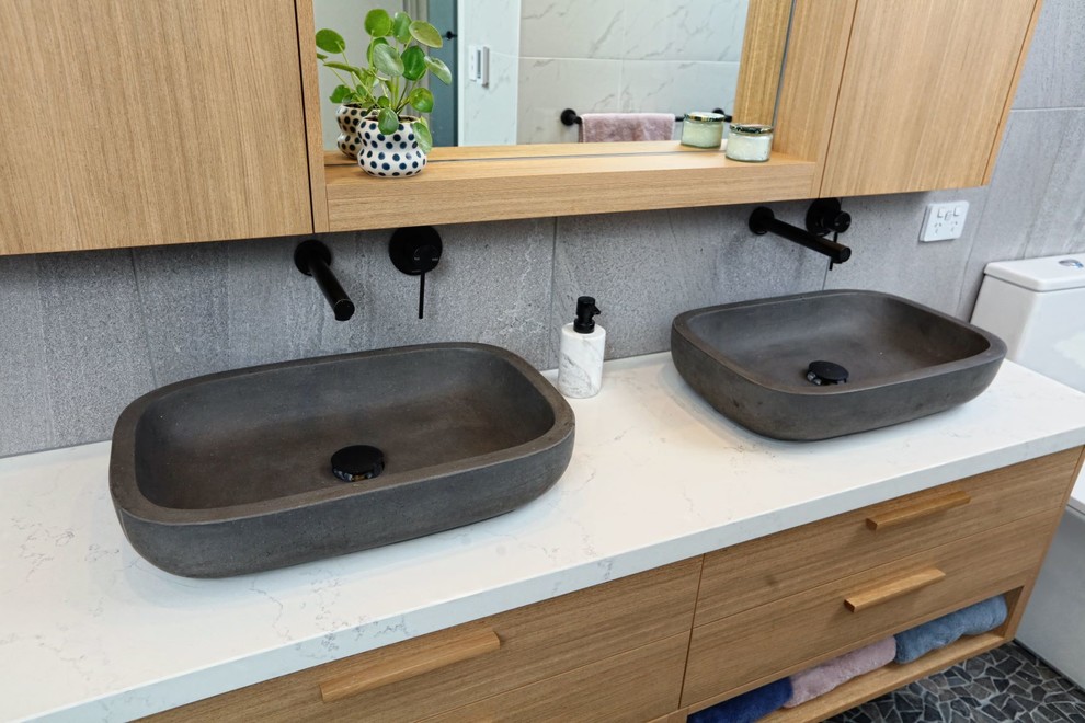 This is an example of a mid-sized contemporary master bathroom in Melbourne with furniture-like cabinets, light wood cabinets, a freestanding tub, a one-piece toilet, porcelain tile, pebble tile floors, a vessel sink, engineered quartz benchtops and multi-coloured floor.