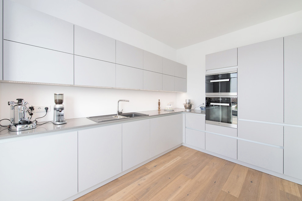 Geschlossene, Große Moderne grifflose Küche ohne Insel in L-Form mit Einbauwaschbecken, flächenbündigen Schrankfronten, grauen Schränken, Quarzwerkstein-Arbeitsplatte, hellem Holzboden und Küchengeräten aus Edelstahl in Stuttgart