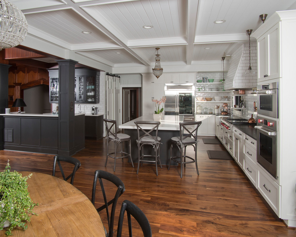 Example of a large transitional u-shaped medium tone wood floor and brown floor eat-in kitchen design with white cabinets, soapstone countertops, white backsplash, ceramic backsplash, stainless steel appliances, an island, a farmhouse sink and shaker cabinets
