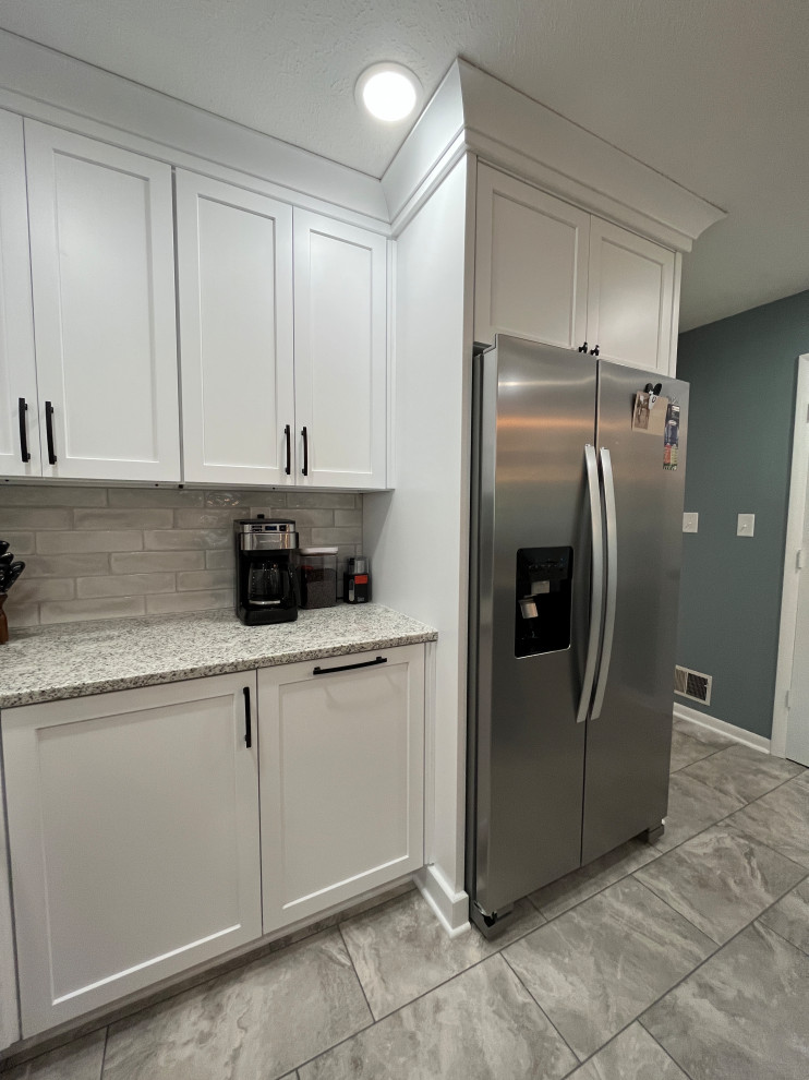 Photo of a medium sized rural l-shaped kitchen/diner in Indianapolis with a submerged sink, shaker cabinets, white cabinets, granite worktops, grey splashback, porcelain splashback, stainless steel appliances, porcelain flooring, an island, grey floors and grey worktops.