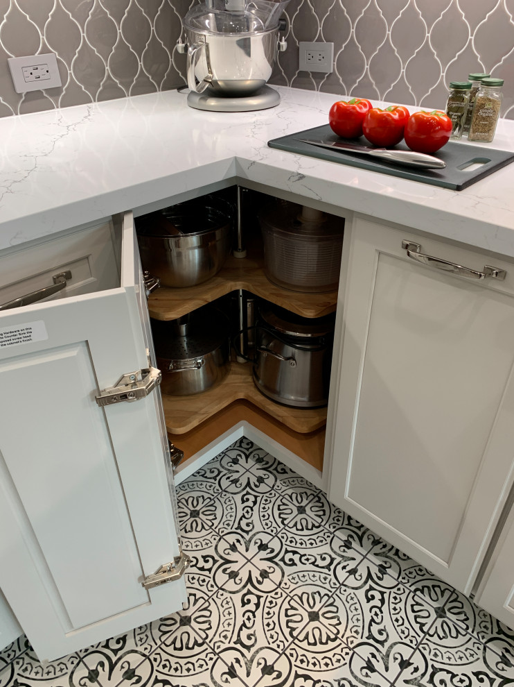 Example of a transitional l-shaped porcelain tile and multicolored floor kitchen design in Houston with a farmhouse sink, recessed-panel cabinets, white cabinets, quartz countertops, gray backsplash, ceramic backsplash, stainless steel appliances, an island and white countertops