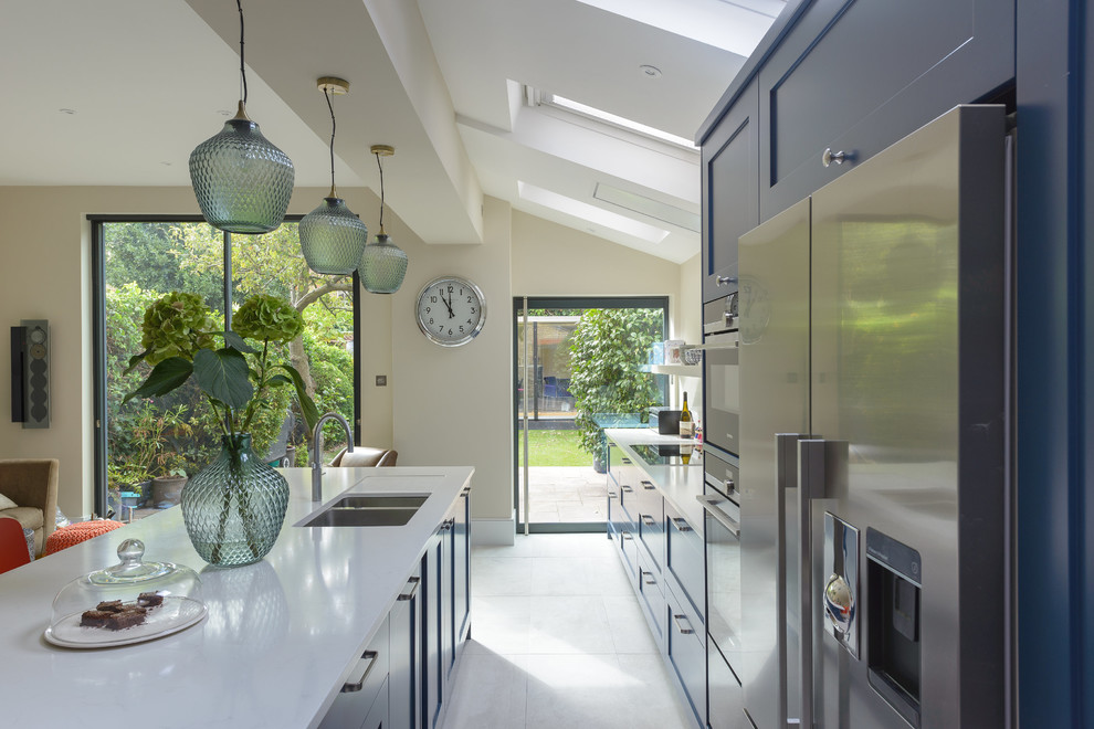 Design ideas for a large contemporary galley eat-in kitchen in London with a farmhouse sink, recessed-panel cabinets, blue cabinets, solid surface benchtops, white splashback, marble splashback, stainless steel appliances, ceramic floors, with island and beige floor.