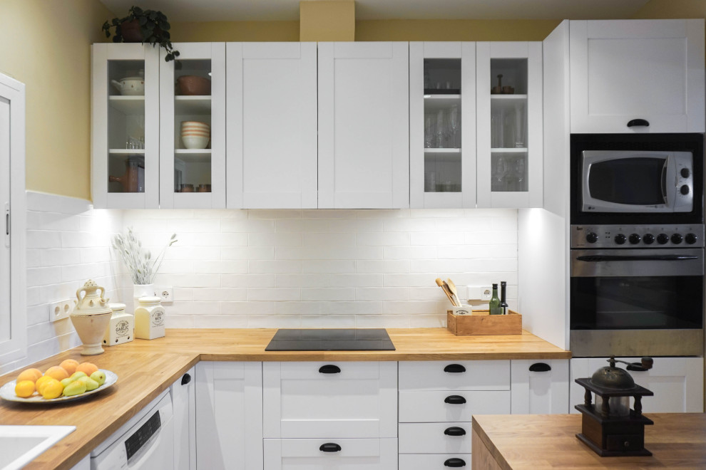 Photo of a mid-sized country u-shaped separate kitchen in Barcelona with a double-bowl sink, glass-front cabinets, white cabinets, wood benchtops, white splashback, ceramic splashback, white appliances, ceramic floors, a peninsula, beige floor and brown benchtop.