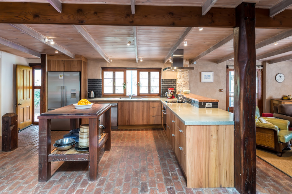 Photo of a mid-sized country l-shaped eat-in kitchen in Adelaide with a double-bowl sink, flat-panel cabinets, medium wood cabinets, concrete benchtops, black splashback, subway tile splashback, stainless steel appliances, brick floors and with island.