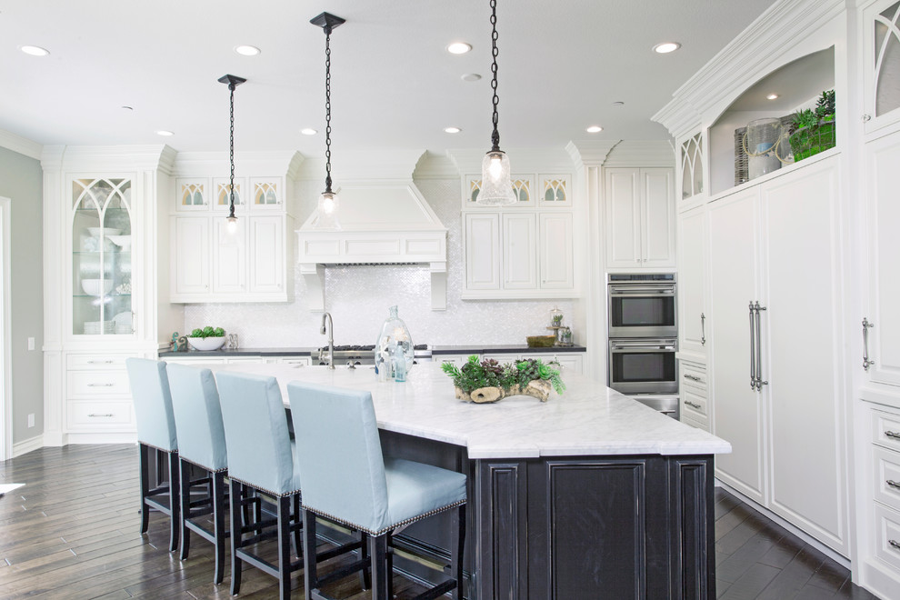 Photo of a beach style l-shaped eat-in kitchen in Orange County with a farmhouse sink, raised-panel cabinets, white cabinets, marble benchtops, white splashback, mosaic tile splashback, stainless steel appliances, dark hardwood floors, with island and brown floor.