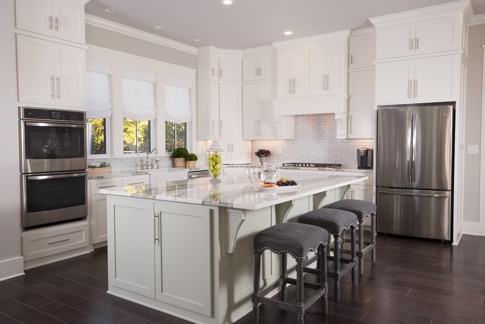 Design ideas for a transitional l-shaped kitchen in Other with a farmhouse sink, shaker cabinets, white cabinets, white splashback, subway tile splashback, stainless steel appliances, dark hardwood floors, with island, brown floor and white benchtop.