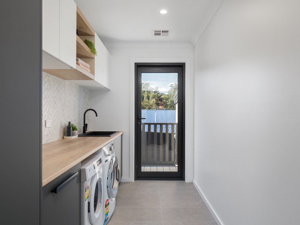 Contemporary galley dedicated laundry room in Hobart with a drop-in sink, grey cabinets, wood benchtops, subway tile splashback, white walls, ceramic floors, a side-by-side washer and dryer and grey floor.