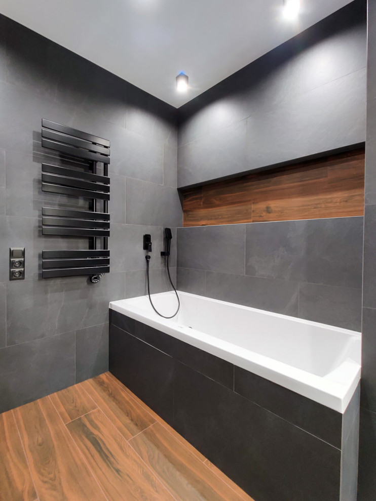 Idéer för att renovera ett litet funkis grå grått en-suite badrum, med släta luckor, vita skåp, ett fristående badkar, en vägghängd toalettstol, grå kakel, grå väggar, ett väggmonterat handfat, kaklad bänkskiva, brunt golv och keramikplattor