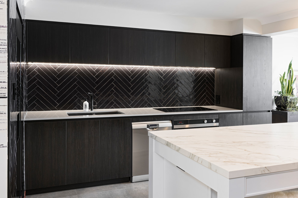 This is an example of a mid-sized contemporary galley open plan kitchen in Perth with an undermount sink, flat-panel cabinets, dark wood cabinets, tile benchtops, black splashback, subway tile splashback, black appliances, cement tiles, with island, grey floor and white benchtop.