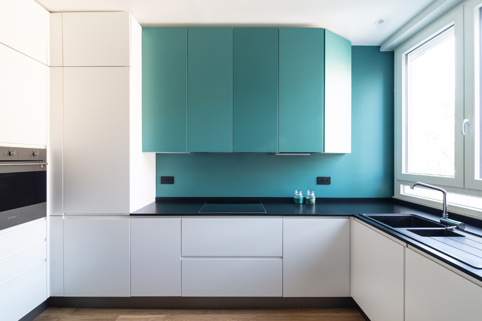 Design ideas for a contemporary l-shaped kitchen in Cagliari with a double-bowl sink, flat-panel cabinets, turquoise cabinets, blue splashback, stainless steel appliances, light hardwood floors, no island, beige floor and black benchtop.