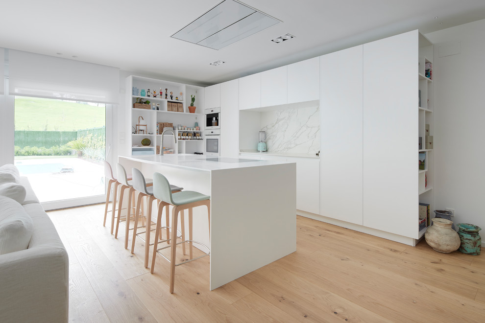 Design ideas for a large contemporary open plan kitchen in Other with an integrated sink, flat-panel cabinets, white cabinets, white splashback, light hardwood floors, with island, white benchtop, stone slab splashback and white appliances.