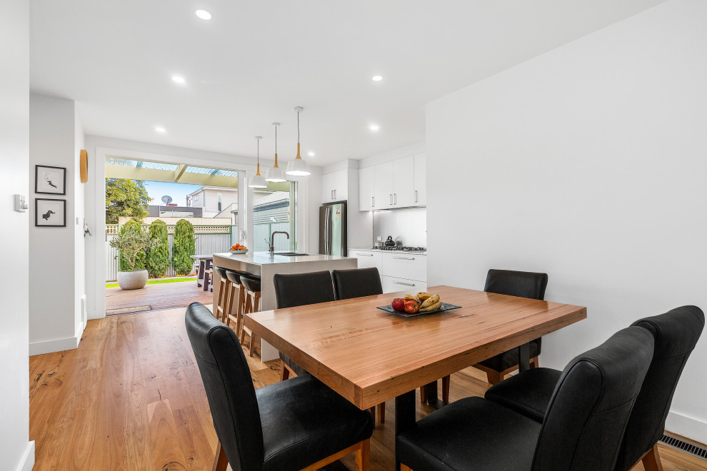 Photo of a mid-sized contemporary galley eat-in kitchen in Melbourne with a double-bowl sink, flat-panel cabinets, white cabinets, quartz benchtops, metallic splashback, glass sheet splashback, stainless steel appliances, medium hardwood floors, with island, brown floor and white benchtop.