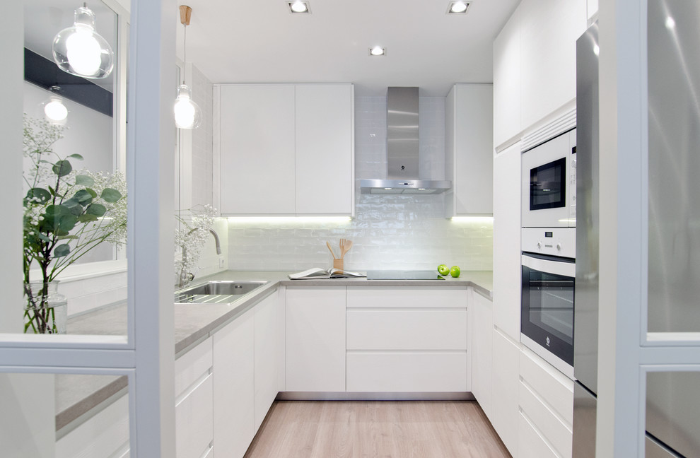 This is an example of a mid-sized contemporary u-shaped separate kitchen in Madrid with a single-bowl sink, white cabinets, concrete benchtops, white splashback, ceramic splashback, white appliances, light hardwood floors, no island, flat-panel cabinets and beige floor.