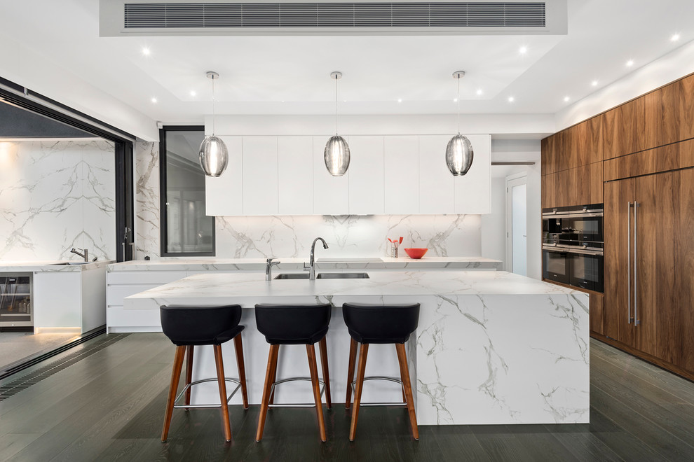 Large contemporary galley open plan kitchen in Melbourne with an integrated sink, flat-panel cabinets, white cabinets, marble benchtops, white splashback, stone slab splashback, black appliances, dark hardwood floors and with island.