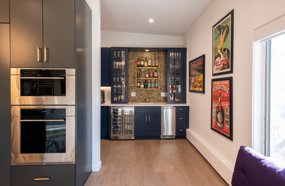 Inspiration for a small modern single-wall wet bar in Denver with an undermount sink, glass-front cabinets, blue cabinets, quartz benchtops, black splashback, light hardwood floors, grey floor and white benchtop.