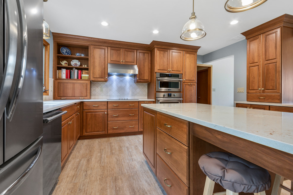 This is an example of a mid-sized traditional l-shaped eat-in kitchen in Milwaukee with an undermount sink, raised-panel cabinets, brown cabinets, quartz benchtops, beige splashback, stone tile splashback, stainless steel appliances, vinyl floors, with island, beige floor and white benchtop.