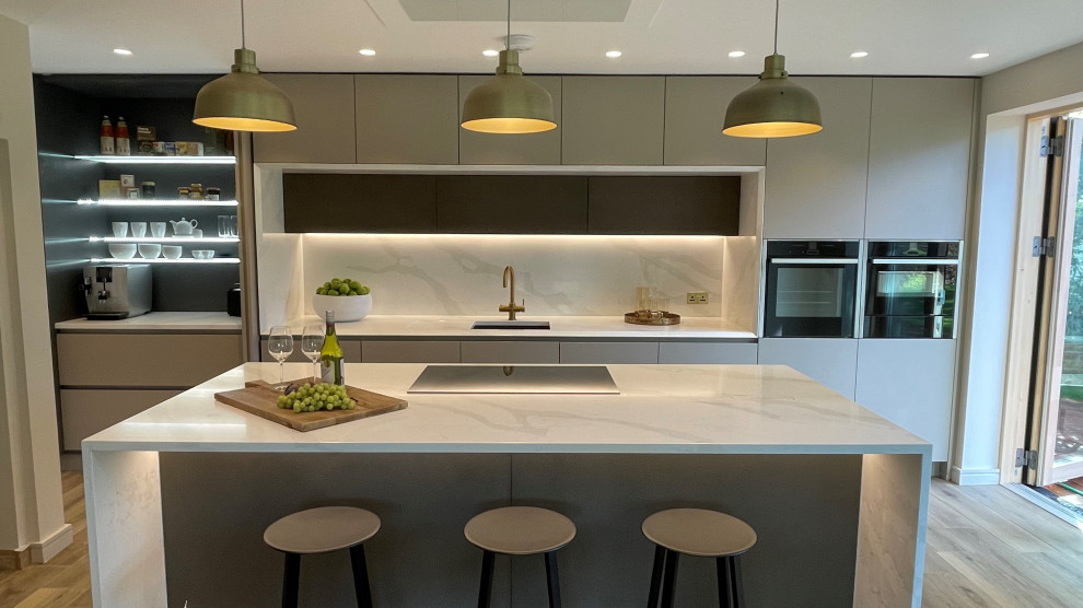 Photo of a large contemporary galley open plan kitchen in Manchester with an undermount sink, flat-panel cabinets, beige cabinets, quartzite benchtops, white splashback, marble splashback, black appliances, medium hardwood floors, with island, brown floor, white benchtop and coffered.