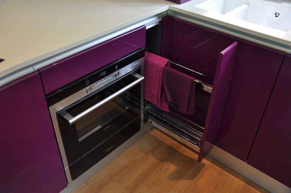 Inspiration for a mid-sized contemporary u-shaped open plan kitchen in Kent with a drop-in sink, flat-panel cabinets, purple cabinets, quartzite benchtops, glass sheet splashback, black appliances, light hardwood floors, a peninsula, brown floor and white benchtop.