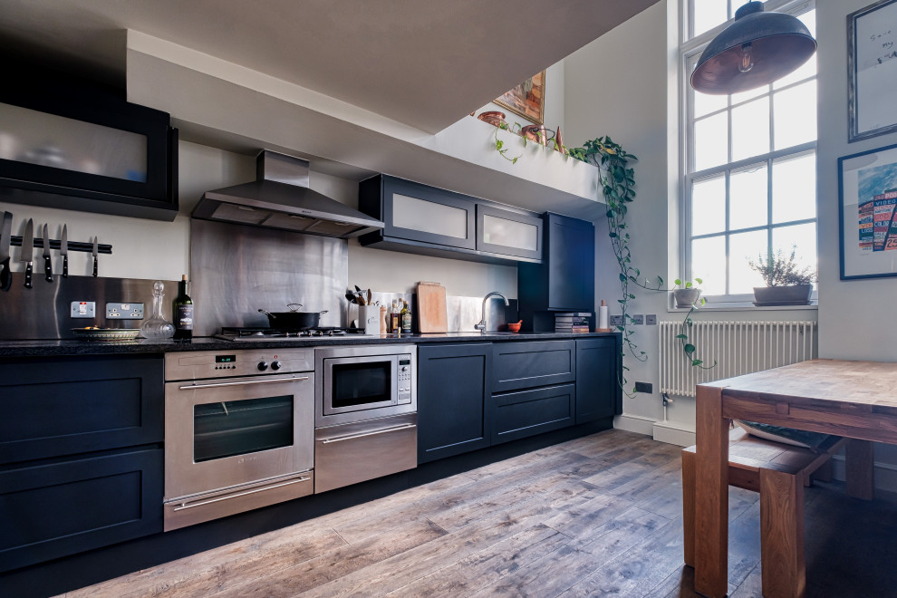 Inspiration for a mid-sized industrial single-wall eat-in kitchen in London with an integrated sink, shaker cabinets, black cabinets, granite benchtops, metallic splashback, metal splashback, stainless steel appliances, dark hardwood floors, brown floor, black benchtop and vaulted.